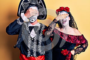 Young couple wearing mexican day of the dead costume over yellow smiling and laughing with hand on face covering eyes for surprise