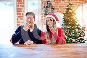 Young couple wearing santa claus hat sitting on chair and table around christmas tree at home bored yawning tired covering mouth