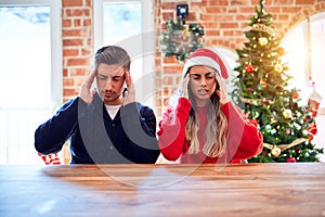 Young couple wearing santa claus hat sitting on chair and table around christmas tree at home with hand on head for pain in head