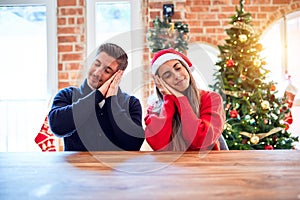 Young couple wearing santa claus hat sitting on chair and table around christmas tree at home sleeping tired dreaming and posing