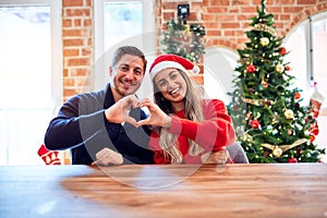 Young couple wearing santa claus hat sitting on chair and table around christmas tree at home smiling in love doing heart symbol