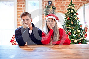 Young couple wearing santa claus hat sitting on chair and table around christmas tree at home thinking looking tired and bored