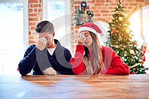 Young couple wearing santa claus hat sitting on chair and table around christmas tree at home tired rubbing nose and eyes feeling