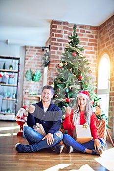 Young couple wearing santa claus hat sitting on the floor around christmas tree at home smiling with happy face looking and