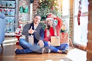 Young couple wearing santa claus hat sitting on the floor around christmas tree at home smiling making frame with hands and