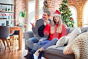 Young couple wearing santa claus hat sitting on the sofa around christmas tree at home smiling with happy face looking and