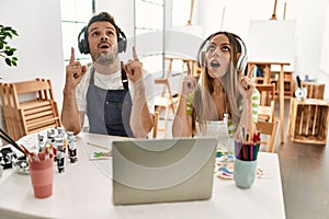 Young couple of wife and husband at art studio looking at video on laptop amazed and surprised looking up and pointing with