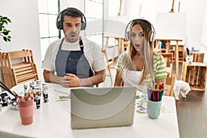 Young couple of wife and husband at art studio looking at video on laptop scared and amazed with open mouth for surprise,