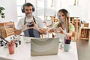 Young couple of wife and husband at art studio looking at video on laptop smiling in love showing heart symbol and shape with
