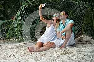 The young couple, the woman and the man sit on the beach and will take the selfies dressed in bright clothes and in sunglasses.