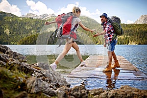 Young couple on wooden jetty by lake. Woman jumping, man lending a hand to her. Summer trip in nature. Lifestyle, togetherness,