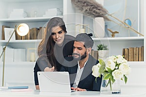 Young couple working from home. Hispanic man in suit using laptop and his girlfirend leaning on him pointing at the