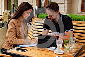 Young Couple Is Working In Outdoor Cafe On Laptop And Smiling.