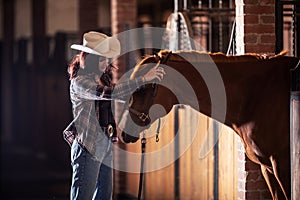 Young cowgirl takes bridle off the horse& x27;s head inside the stable after a ride