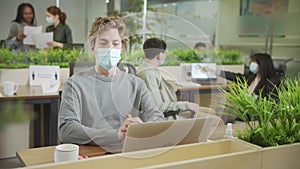 A young coworker is sitting in a facial protective mask and looking to the camera
