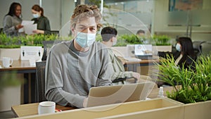 A young coworker is sitting in a facial protective mask and looking to the camera