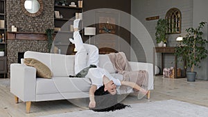 Young crazy funny carefree woman lying upside down on cozy sofa in living room shaking arms and legs dances lying enjoys