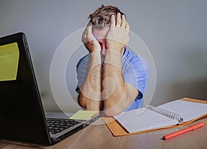 Young crazy stressed and overwhelmed man working messy at office desk desperate with laptop computer covering face with hands frus