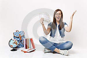 Young crazy woman student screaming spreading hands holding bundle lots of dollars, cash money sit near globe backpack