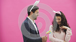 Young creative couple on pink background. With hackneyed ears on the head. During this man gives decorative multicolored