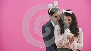 Young creative couple on pink background. With hackneyed ears on the head. During this man gives a soft toy with a hare