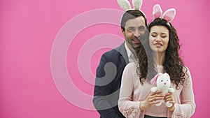 Young creative couple on pink background. With hackneyed ears on the head. During this, a wife holds a soft toy hare, a