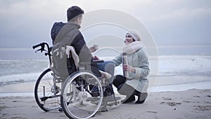 Young crippled Caucasian boy in wheelchair drinking tea on sea shore and talking with beautiful woman sitting on hunkers