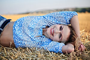 Young curly blond woman, wearing jeans shorts and light blue shirt, lying on bale on field in summer. Close-up female portrait in