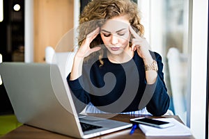 Young curly businesswoman suffering from headache in front of laptop at office desk in modern office