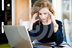 Young curly businesswoman suffering from headache in front of laptop at office desk in modern office