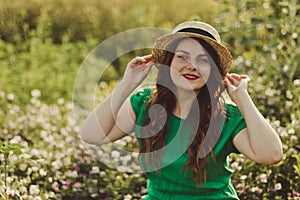 Young curly Caucasian girl in a green dress and a straw hat looks at the camera and smiles in a flower field on a sunny