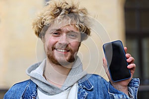 A young curly-haired guy with a beard holds a mobile phone in his hands, mockup