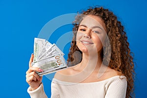 Young curly happy woman holding pile of dollar banknotes against blue background