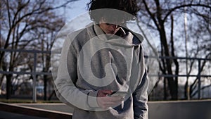 Young curly headed male skateboarder browsing in his mobile phone in the skate park on a sunny day. Lean on railing with