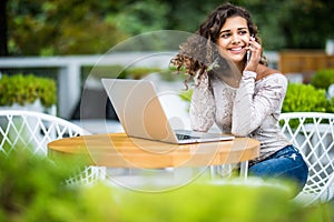 Young curly latin woman calling with cell telephone while sitting alone in street cafe during free time while work on her modern l