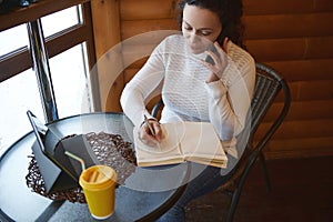 Young curly woman concentrated at work, writing on diary, talking by smartphone while sitting near window in a wooden cafeteria