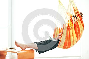 Young curly woman sitting in hammock near big bright window with laptop