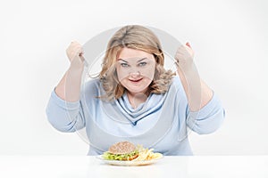 Young curvy fat woman in casual blue clothes on a white background at the table eagerly eating fast food, hamburger and