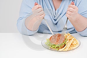 Young curvy fat woman in casual blue clothes on a white background at the table eagerly eating fast food, hamburger and