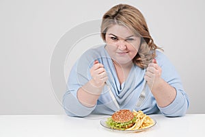 Young curvy fat woman in casual blue clothes on a white background at the table eagerly eating fast food, hamburger and