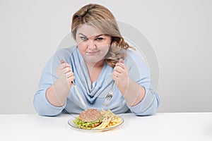 Young curvy fat woman in casual blue clothes on a white background at the table eagerly eating fast food, hamburger and