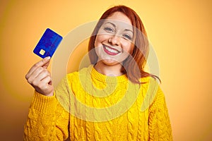 Young customer redhead woman holding credit card over yellow isolated background with a happy face standing and smiling with a