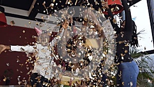 Young cute african woman sitting with gifts box while her friends throwing golden confetti at the birthday party.