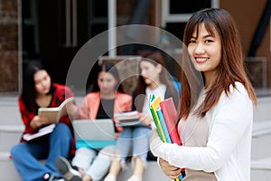 Young and cute Asian college student girls holding books, pose to camera with group of friends blur in background in front of