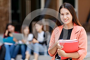 Young and cute Asian college student girls holding books, pose to camera with group of friends blur in background in front of