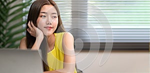 Young cute Asian office woman sitting daydreaming and look sideways to the copy space to the right at the desk in the office,