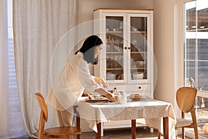 A young cute Asian woman sets the table in the dining room at home. A charming Korean girl sets out plates and cups for a family