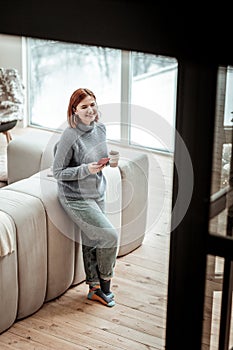 Young cute beaming girl in a grey sweater standing near the sofa