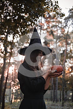 Young cute beautiful woman in dark dress and witch`s hat holds an orange pumpkin in her hands. Halloween party costume