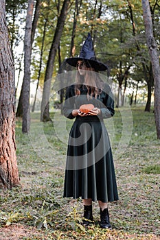 Young cute beautiful woman in dark dress and witch`s hat holds an orange pumpkin in her hands. Halloween party costume